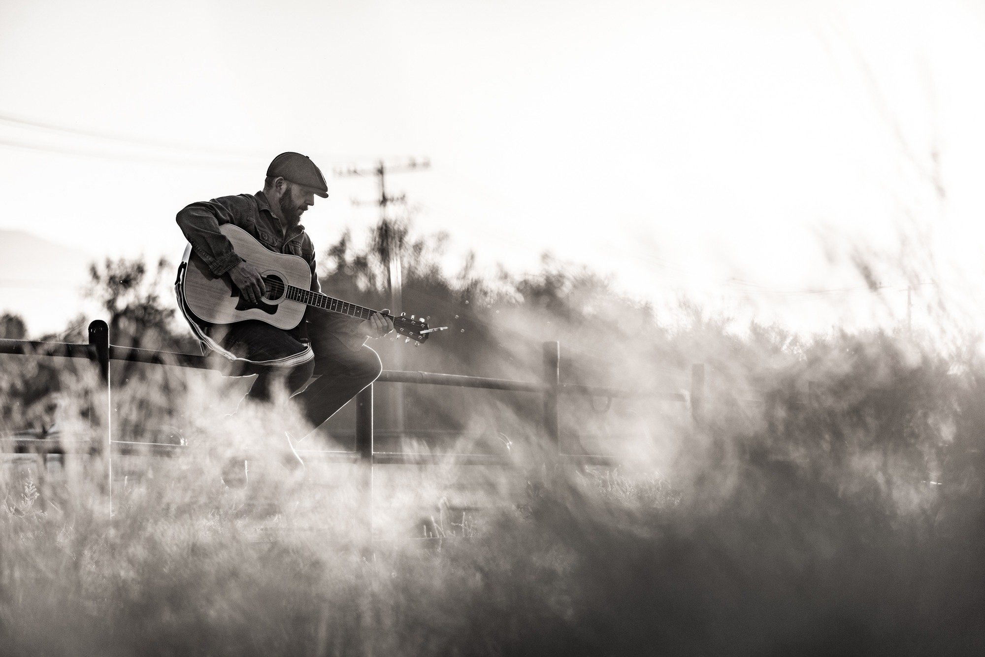 Everett Rogers sitting on a fence rail with an acoustic guitar, backlit in tall grass
