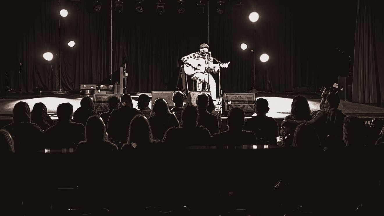 Everett Rogers alone in a stage light, seen over the heads of a seated audience