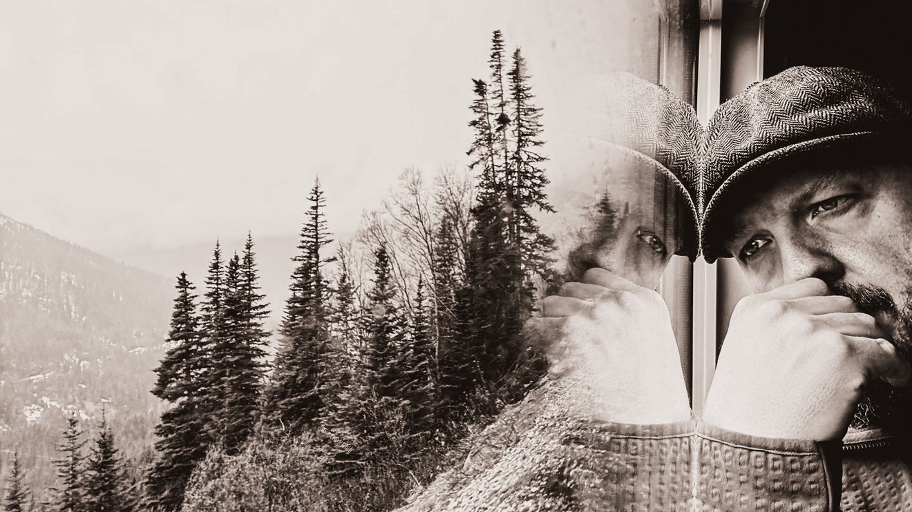 Everett Rogers at a window, pines and mountains carried in the reflection